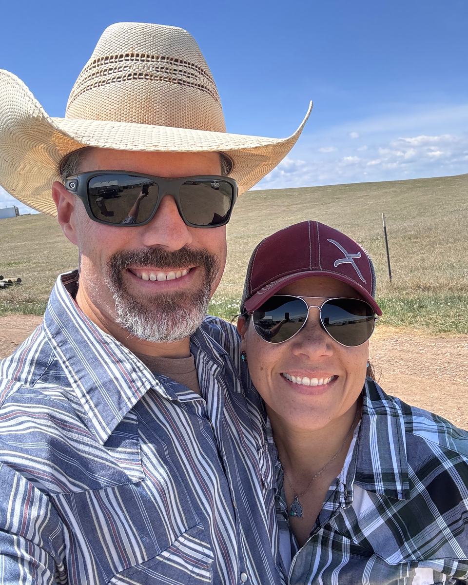 Ed and Samantha smiling together outside on the ranch, in a straw hat and a ball cap, with pasture and blue sky behind them.